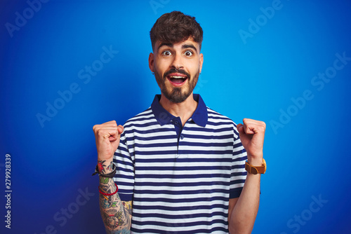 Young man with tattoo wearing striped polo standing over isolated blue background celebrating surprised and amazed for success with arms raised and open eyes. Winner concept.