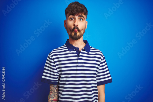 Young man with tattoo wearing striped polo standing over isolated blue background making fish face with lips, crazy and comical gesture. Funny expression.