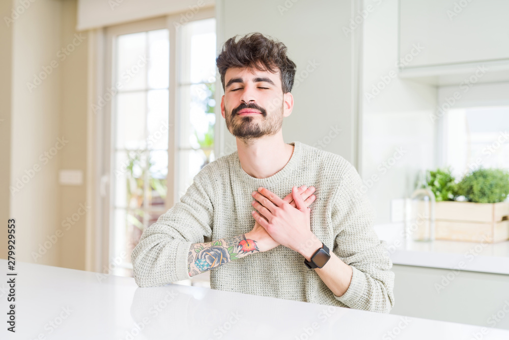 Young man wearing casual sweater sitting on white table smiling with hands on chest with closed eyes and grateful gesture on face. Health concept.