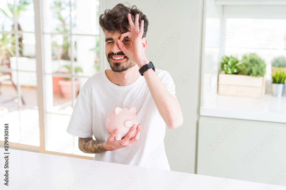 Young man holding piggy bank as insurance investment with happy face smiling doing ok sign with hand on eye looking through fingers