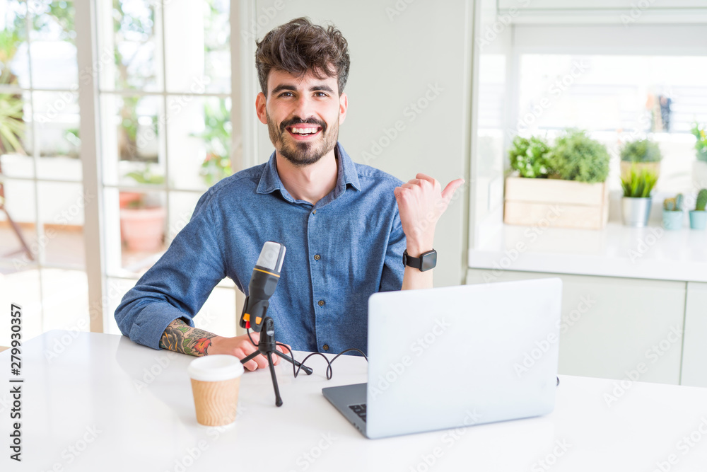 Young man recording podcast using microphone and laptop smiling with happy face looking and pointing to the side with thumb up.
