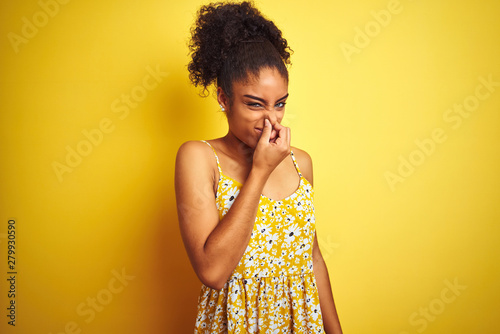 African american woman wearing casual floral dress standing over isolated yellow background smelling something stinky and disgusting, intolerable smell, holding breath with fingers on nose. Bad smells