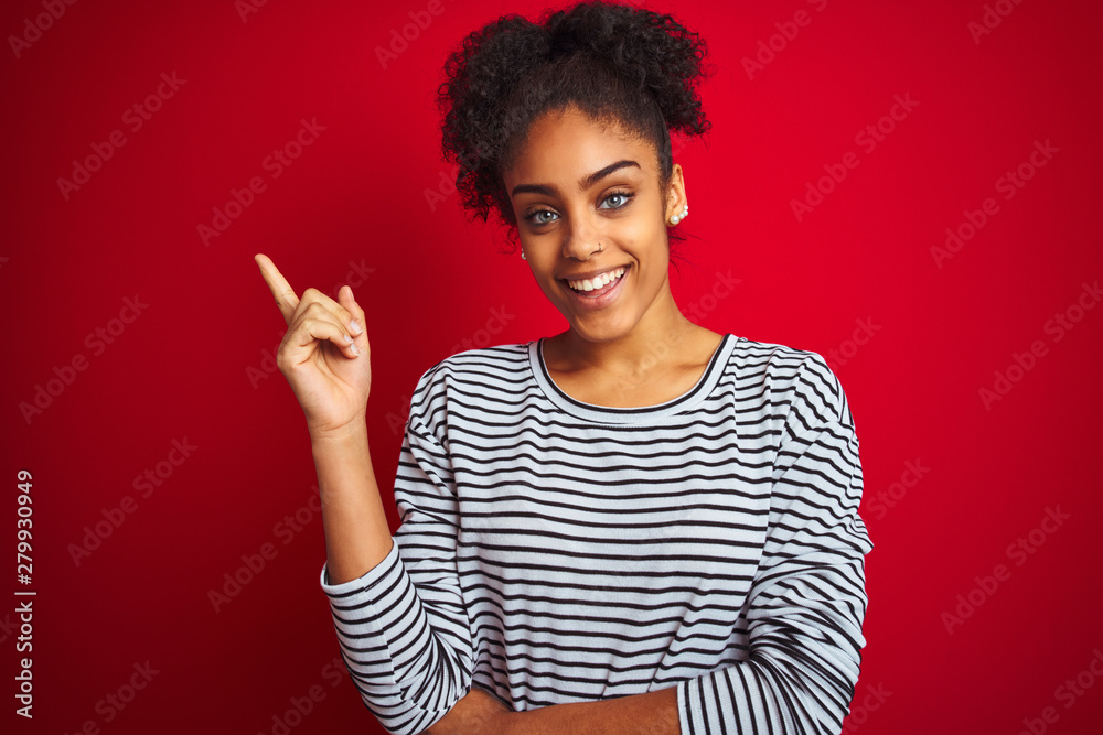 African american woman wearing navy striped t-shirt standing over isolated red background with a big smile on face, pointing with hand and finger to the side looking at the camera. African american woman wearing navy striped t-shirt standing over isolated red background with a big smile on face, pointing with hand and finger to the side looking at the camera.