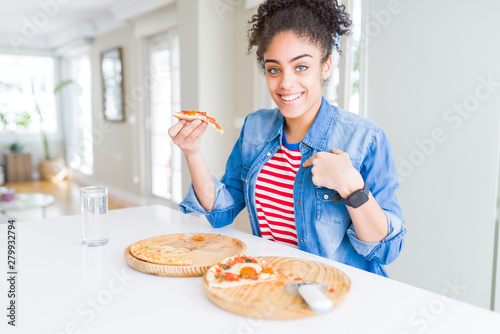 Young african american woman eating two homemade cheese pizzas with surprise face pointing finger to himself
