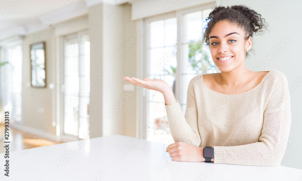 Beautiful young african american woman with afro hair sitting on table at home smiling cheerful presenting and pointing with palm of hand looking at the camera. Beautiful young african american woman with afro hair sitting on table at home smiling cheerful presenting and pointing with palm of hand looking at the camera.