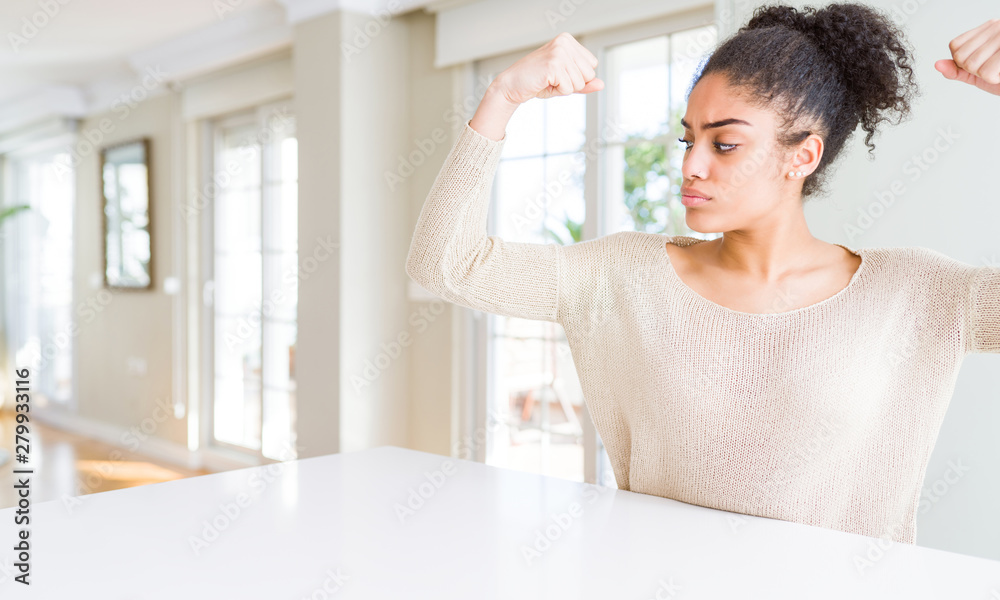 Beautiful young african american woman with afro hair sitting on table at home showing arms muscles smiling proud. Fitness concept. Beautiful young african american woman with afro hair sitting on table at home showing arms muscles smiling proud. Fitness concept.
