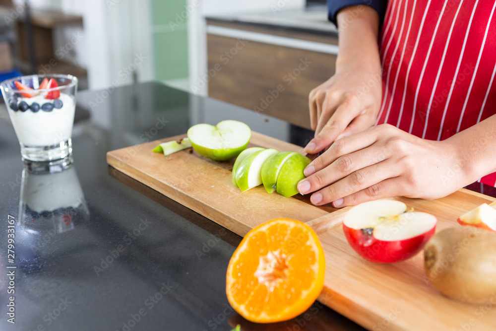 Closeup of young asian man with apron cutting red and green apple kiwi orange on wooden black table of kitchen room