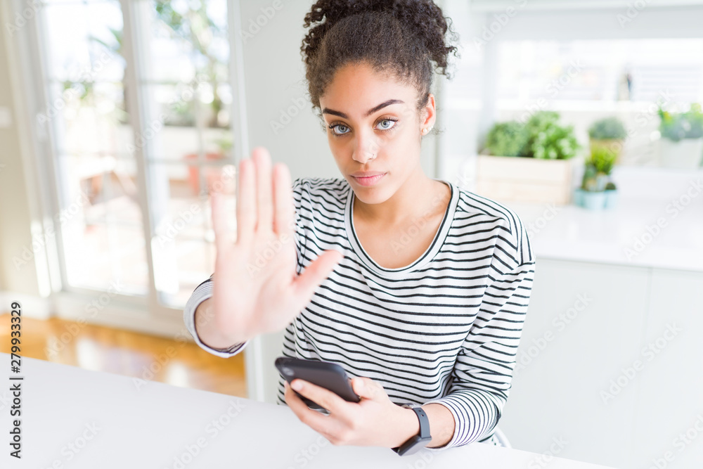 Young african american woman using smartphone texting a message with open hand doing stop sign with serious and confident expression, defense gesture