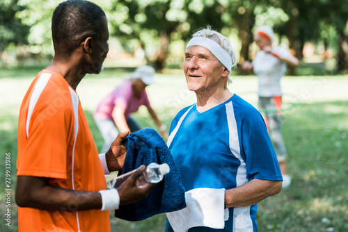 selective focus of happy and sportive senior man holding bottle with water and looking at african american man