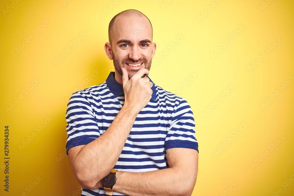 Young bald man with beard wearing casual striped blue t-shirt over yellow isolated background looking confident at the camera smiling with crossed arms and hand raised on chin. Thinking positive. Young bald man with beard wearing casual striped blue t-shirt over yellow isolated background looking confident at the camera smiling with crossed arms and hand raised on chin. Thinking positive.