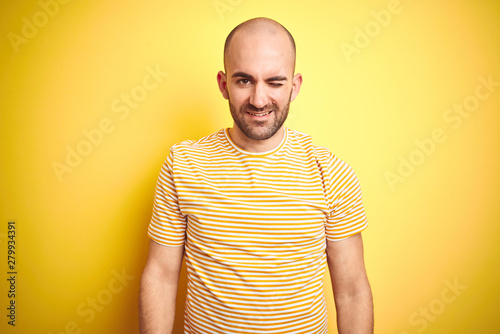 Young bald man with beard wearing casual striped t-shirt over yellow isolated background winking looking at the camera with sexy expression, cheerful and happy face.