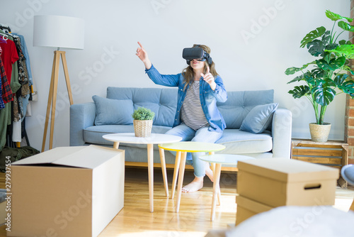 Young blonde woman wearing virtual reality glasses playing a simulation game sitting on the sofa around cardboard boxes moving to a new house