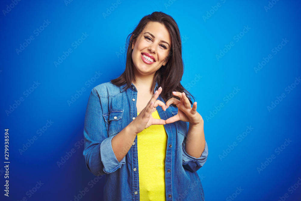 Young beautiful brunette woman wearing casual blue denim jacket over blue isolated background smiling in love showing heart symbol and shape with hands. Romantic concept. Young beautiful brunette woman wearing casual blue denim jacket over blue isolated background smiling in love showing heart symbol and shape with hands. Romantic concept.