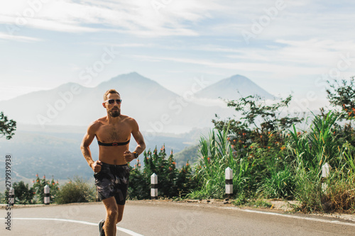 Man running topless in uphill on the asphalt road in hot summer weather. Panoramic mountain view on background. Using chest heart rate monitor.