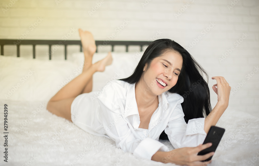 Beautiful young woman in white shirt using video call or self portrait selfie from smartphone on white bed in bedroom, Happy woman relaxing, Smiles and laughs, Communication and social network-Image
