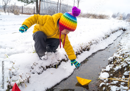 child reaches for a paper boat along a stream in winter