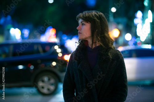Caucasian pedestrian woman walking the street at night in the city with moving cars in the background.  She looks nervous or unsure like a lost tourist or afraid of commuting alone.