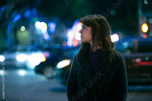 Caucasian pedestrian woman walking the street at night in the city with moving cars in the background.  She looks nervous or unsure like a lost tourist or afraid of commuting alone.