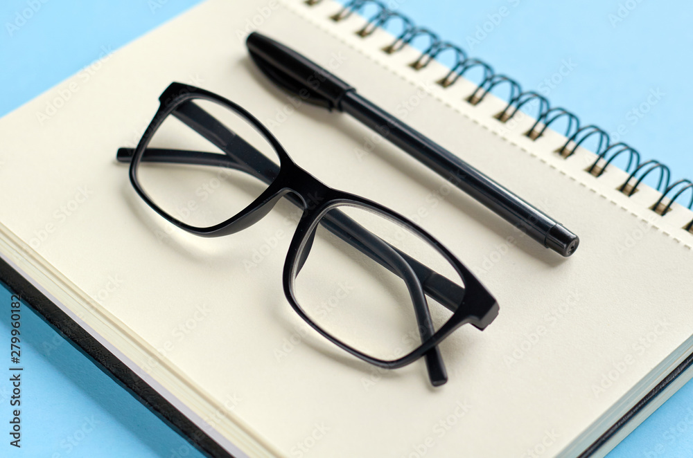 Black glasses, pen and paper notebook on blue background composition. Black glasses, pen and paper notebook on blue background composition.