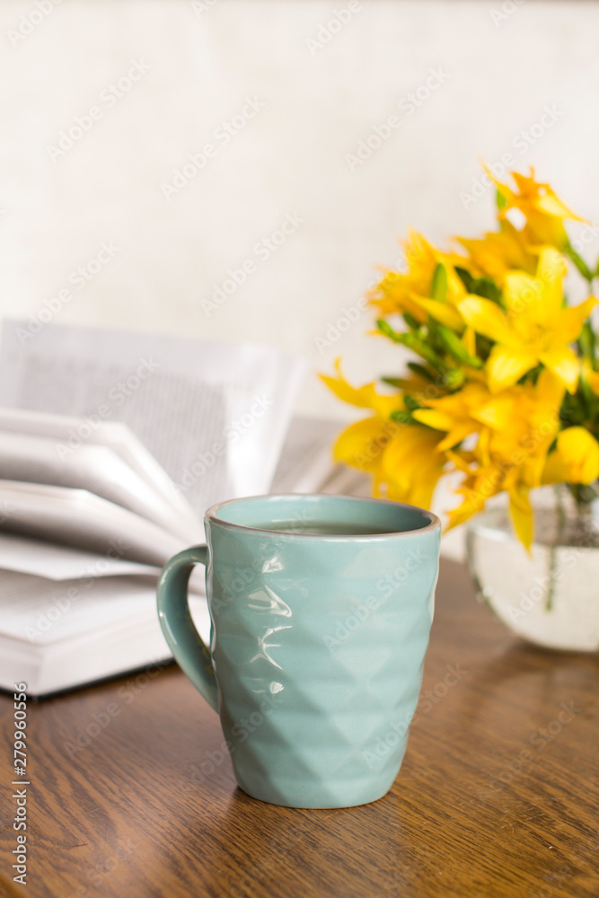 blue coffee mug, yellow flowers in transparent bowl and a book on a wooden table
