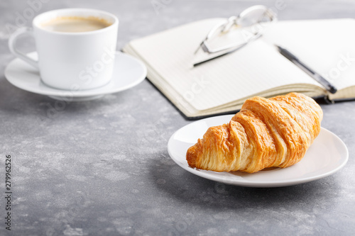 Morning cup of coffee and croissant for breakfast. Day planning, empty notebook, pen and glasses on gray table.