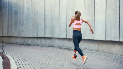 On the way to success.Back view of young disabled woman with leg prosthesis in comfortable sports clothing is running outdoors along the street