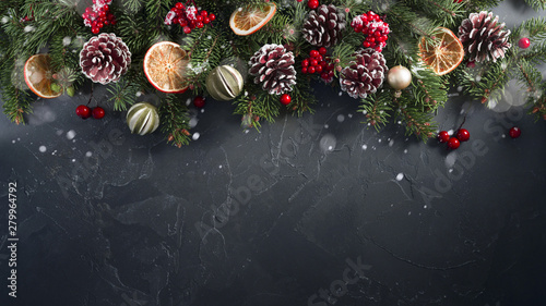 Festive Christmas greeting layout template with fir branches decorated with natural dried decor of oranges, lemons, fir cones and red berries. Dark black textured background, copy space.