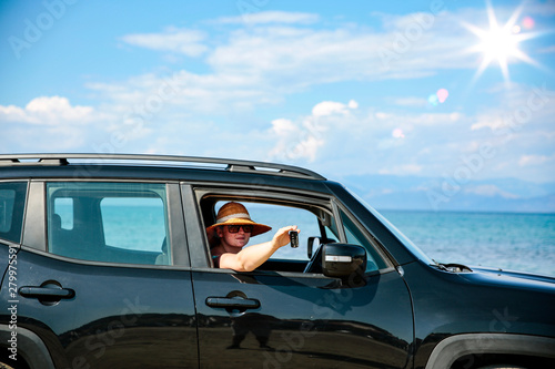 A woman wearing sunglasses and a hat sitting in a summer black car on sandy beach view.