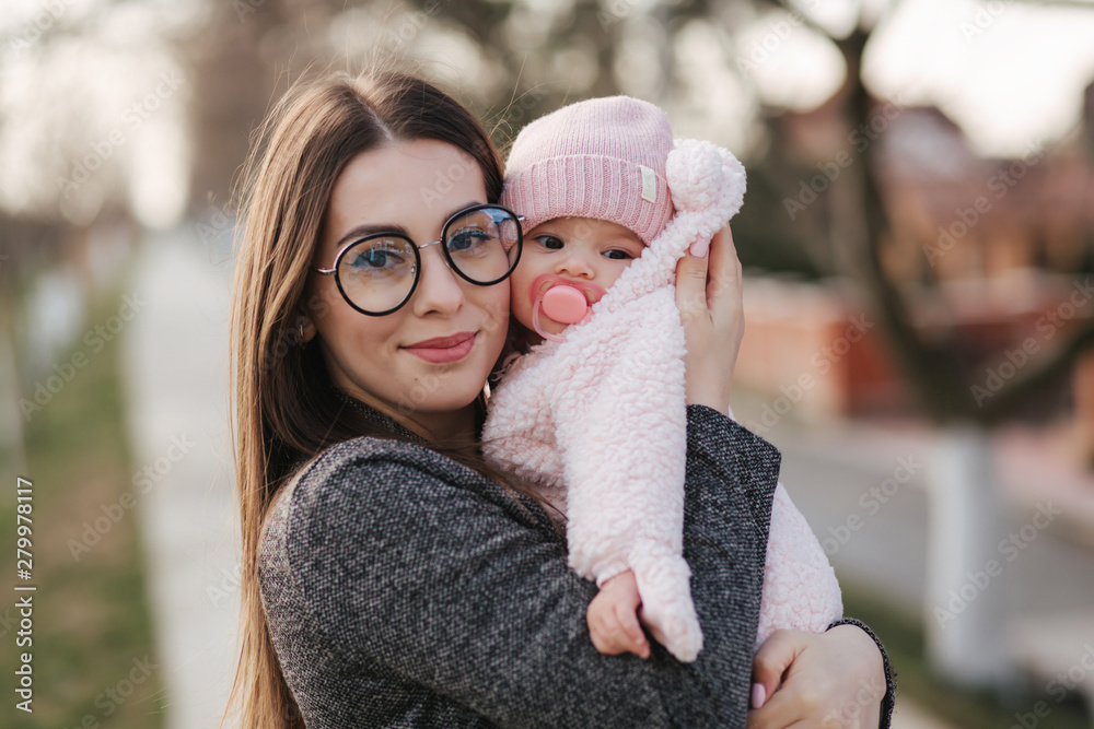 Portrait of mother and her little baby girl. Beautiful mom and cute baby. Mother hud her daughter. Baby dressed in peanch colour hat and nipple toy Portrait of mother and her little baby girl. Beautiful mom and cute baby. Mother hud her daughter. Baby dressed in peanch colour hat and nipple toy