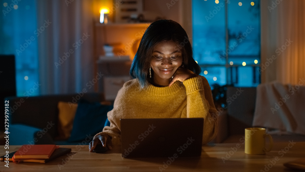 Portrait of Beautiful Black Girl Uses Computer while Sitting at Her Desk at Home, She's Wearing Warm Sweater. In the Evening Creative Woman Works on a Computer In Her Cozy Living Room.  - obrazy, fototapety, plakaty 