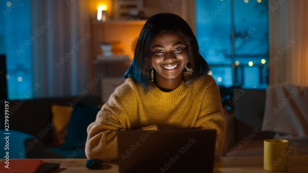 Portrait of Beautiful Black Girl Uses Computer while Sitting at Her Desk at Home, She's Wearing Warm Sweater. In the Evening Creative Woman Works on a Computer In Her Cozy Living Room. Portrait of Beautiful Black Girl Uses Computer while Sitting at Her Desk at Home, She's Wearing Warm Sweater. In the Evening Creative Woman Works on a Computer In Her Cozy Living Room.