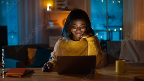 Portrait of Beautiful Black Girl Uses Computer while Sitting at Her Desk at Home, She's Wearing Warm Sweater. In the Evening Creative Woman Works on a Computer In Her Cozy Living Room. 