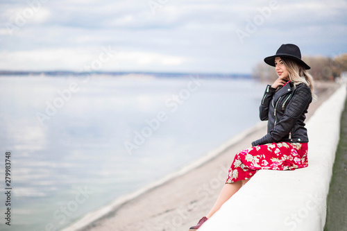 Beautiful girl in a red dress and black hat sits on the waterfront