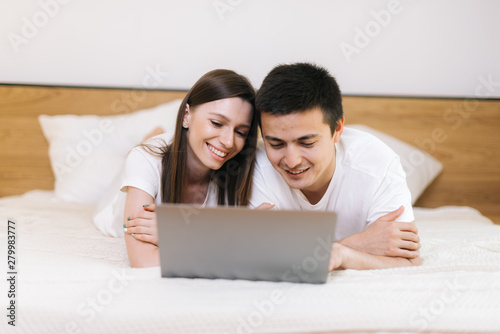 Young couple relaxing in light bed with laptop. Lying together in light bedroom. Happy smiling young couple in love at home, laying in bed, looking at laptop computer © dikushin