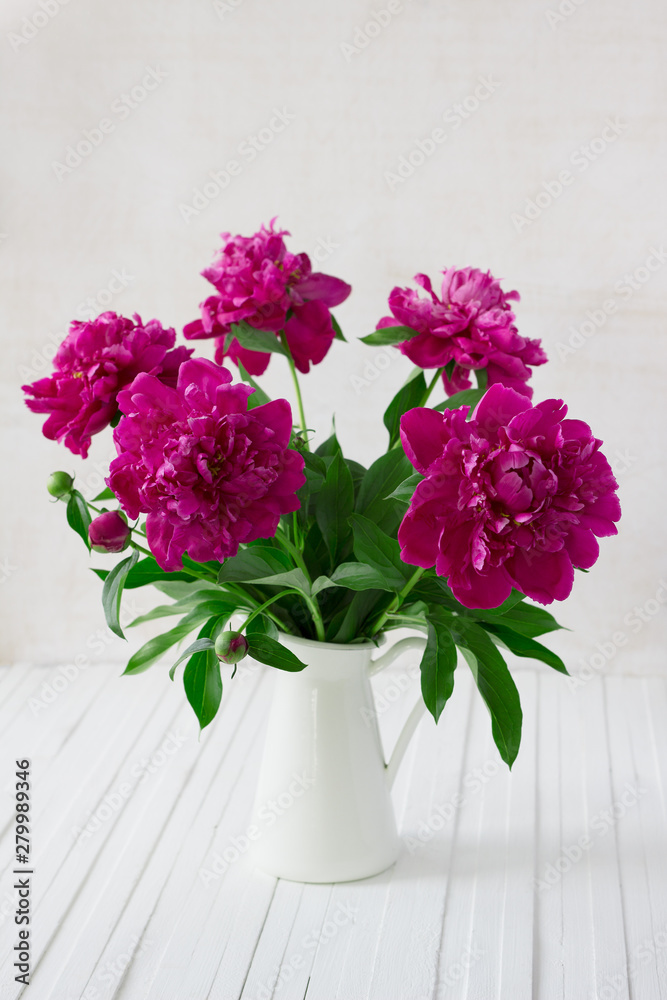 Purple garden peonies in a white enamel jug on a white wooden background, rustic style