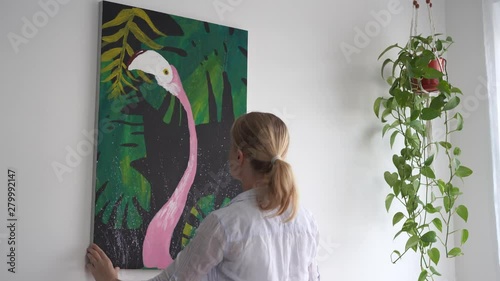 Moving to a new home. A young blonde woman laying down a picture on canvas in a room.