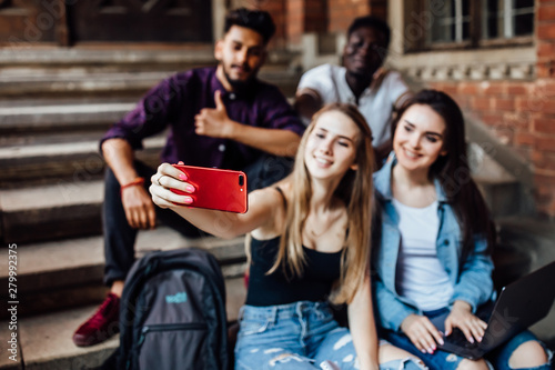 Focus at phone. Young blonde woman making selfie with her friends students, while they are sitting on stairs.