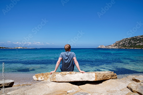 Man sitting on a rock, relaxing and enjoying the beautiful sea view at a beach with a clear blue sky in Sardinia (Italy)