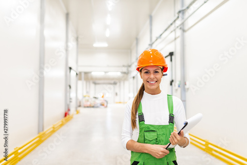 Portrait of a beautiful young female employee in protective uniform holding clipboard, smiling at camera.