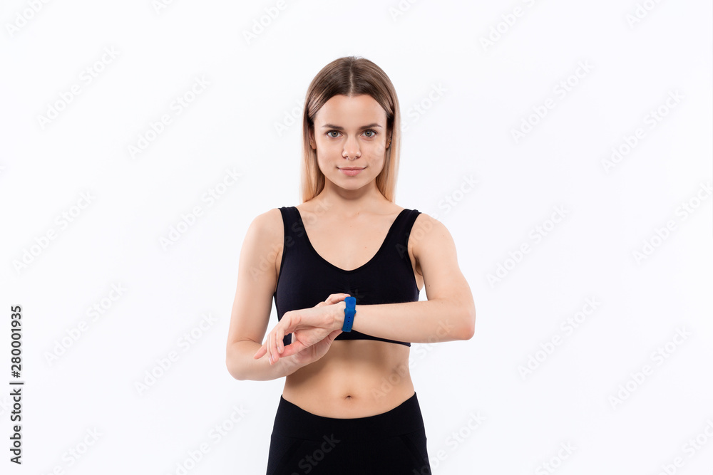 Young sporty blond woman in a black sportswear looking at smart watches checking pulse after workout standing over white background. Young sporty blond woman in a black sportswear looking at smart watches checking pulse after workout standing over white background.