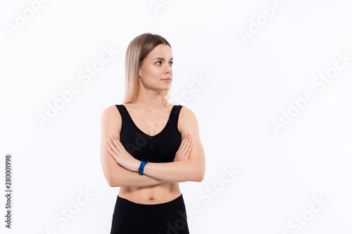 Young sporty blond woman in a black sportswear with smart watches for pulse measuring keeping hands crossed standing over white background.