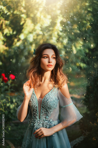 tender girl with cute doll fair face and short dark hair, princess in light blue dress with open shoulders, elegant lady in garden with green trees and red roses on background, art portrait.