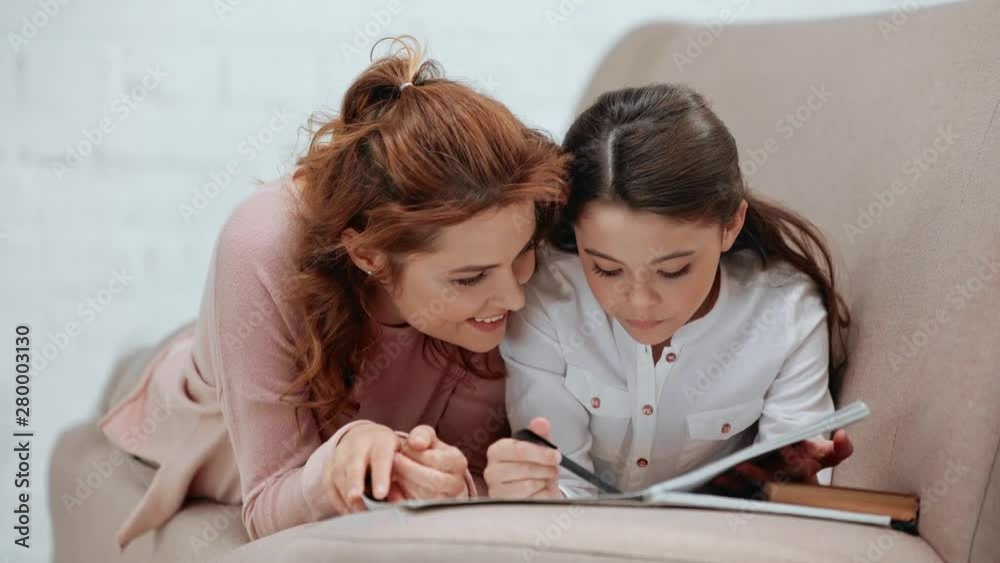 preteen schoolgirl with pen turning pages in copy book and talking to mother, while lying on sofa and doing homework preteen schoolgirl with pen turning pages in copy book and talking to mother, while lying on sofa and doing homework