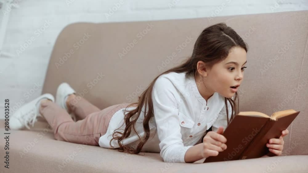 preteen kid reading book, expressing amazement and smiling while lying on sofa at home preteen kid reading book, expressing amazement and smiling while lying on sofa at home