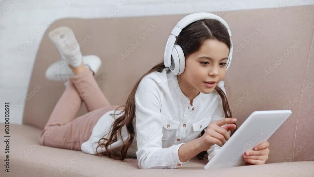 cheerful preteen child lying on sofa, singing while listening music in headphones and using digital tablet cheerful preteen child lying on sofa, singing while listening music in headphones and using digital tablet