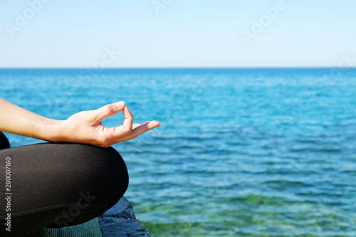 Attractive sporty young yogi woman wearing sportswear practicing yoga, sitting in padmasana lotus pose, meditating on cliff over the calm ocean background. Close up, copy space.