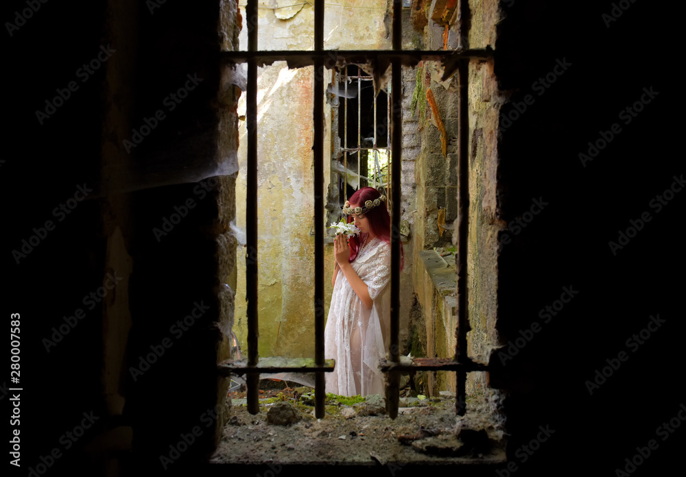A young woman is trapped inside a jail ruin. She is  dressed in white and wears a thorn wreath on her  head. 