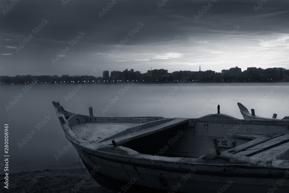 Black and white shot of fishing boat on the beach at sunrise with Egyptian Alexandria city skyline in far distance and cloudysky at sunrise