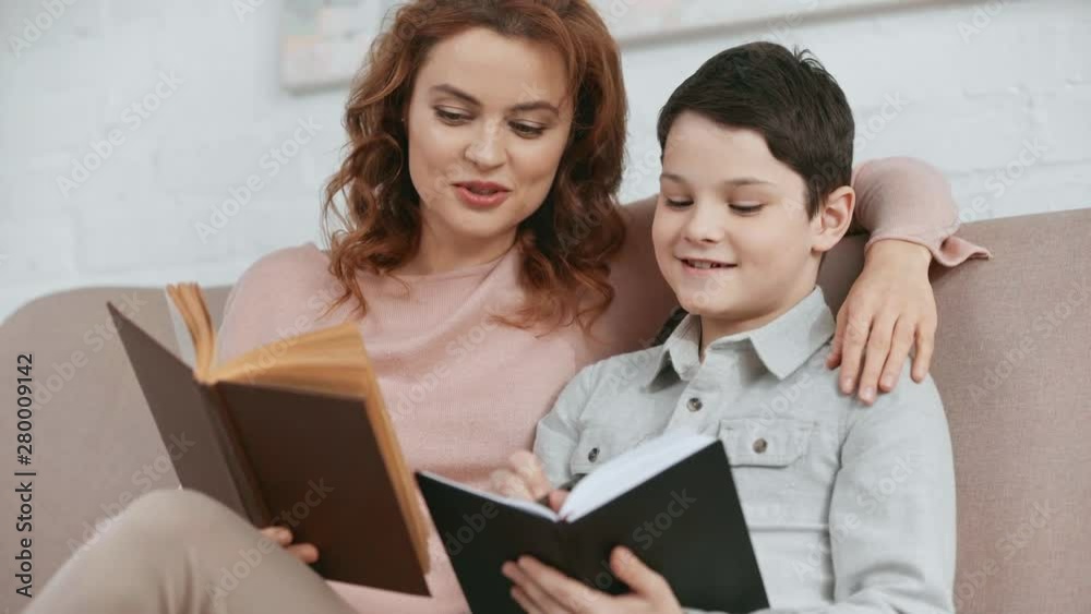 smiling mother sitting on sofa, embracing son and reading book while he writing in textbook smiling mother sitting on sofa, embracing son and reading book while he writing in textbook