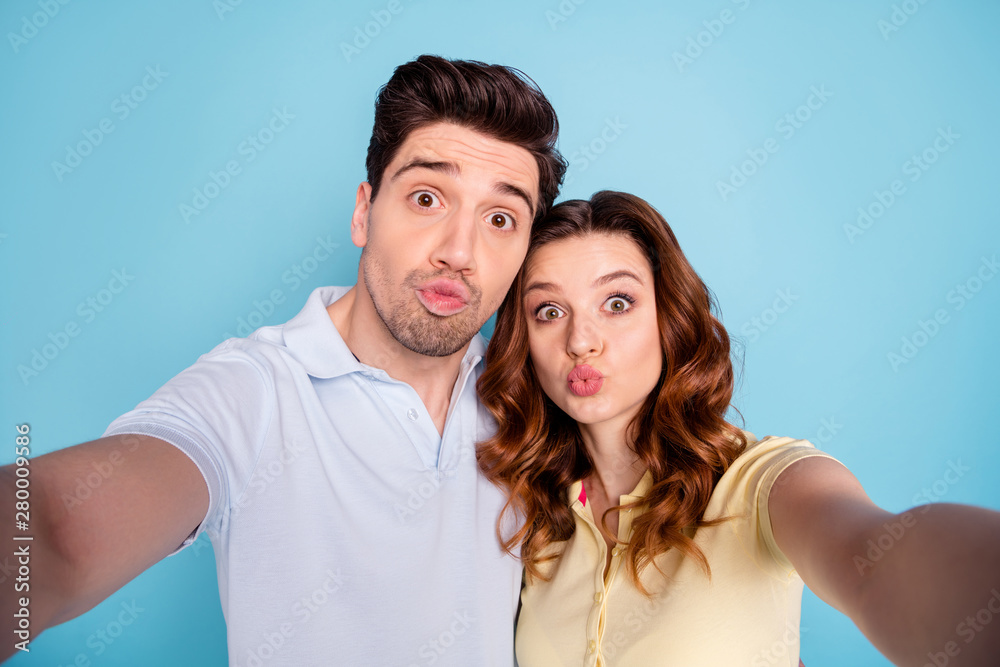Close up photo of pretty foxy lady brunet guy pair making selfies send air kiss followers wear casual t-shirts isolated blue background Close up photo of pretty foxy lady brunet guy pair making selfies send air kiss followers wear casual t-shirts isolated blue background